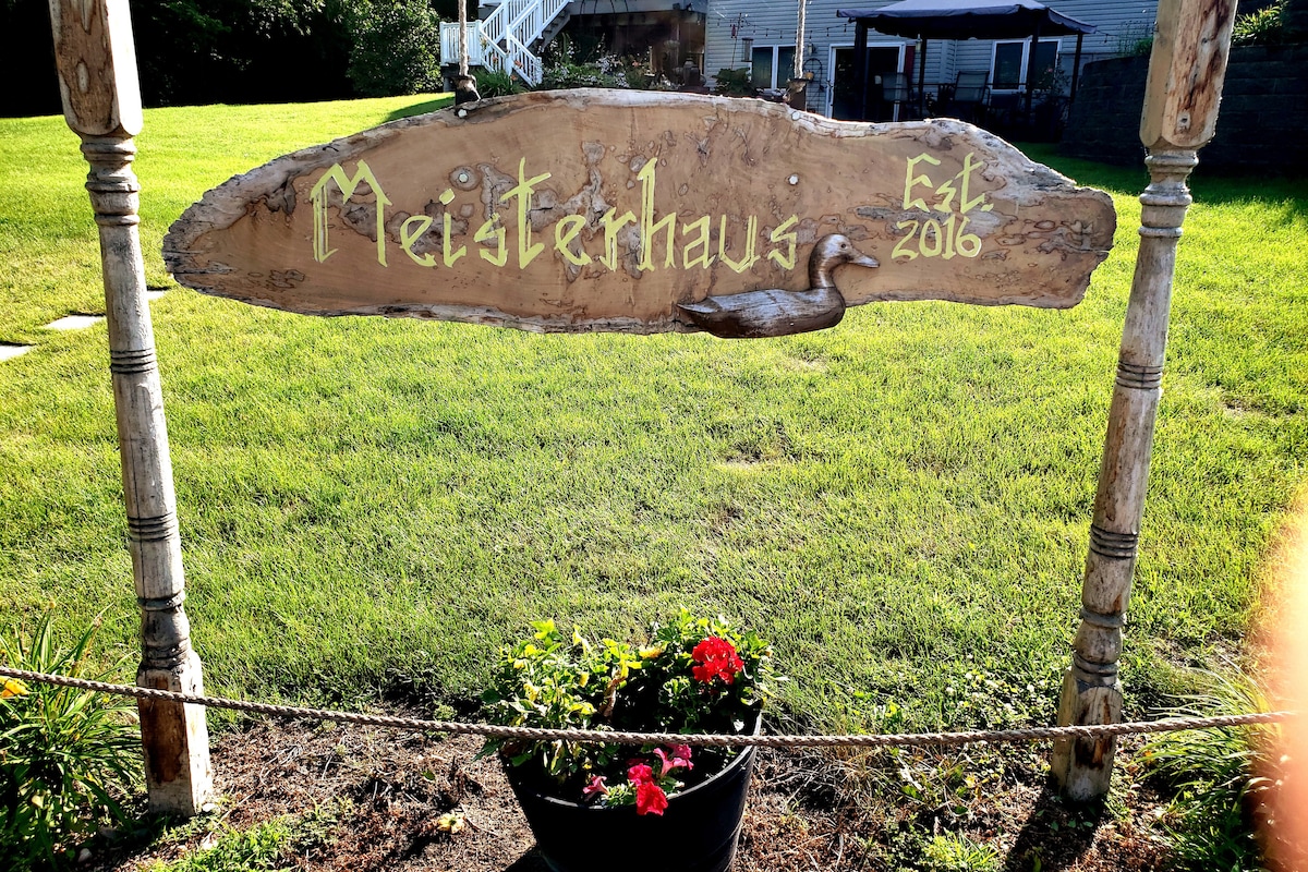 A wooden sign featuring the name 'Meisterhaus' with an engraved duck design sits prominently on a grassy area. Below the sign, a black planter holds vibrant flowers, contributing to the welcoming entrance of the property.