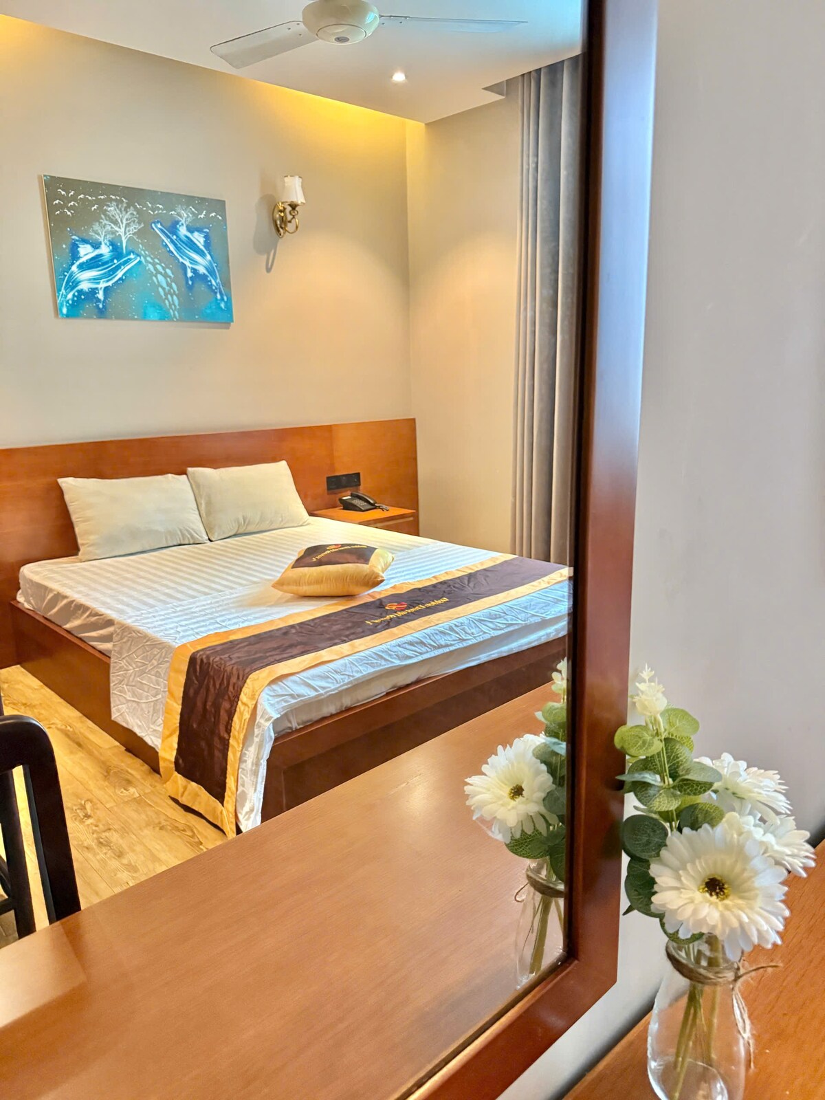 A bright and inviting bedroom features a spacious bed with neutral bedding accented by a decorative throw. Natural light filters through curtains, enhancing the serene environment. A mirror reflects the room's decor, which includes a wall art piece and a vase of flowers on the adjacent table.