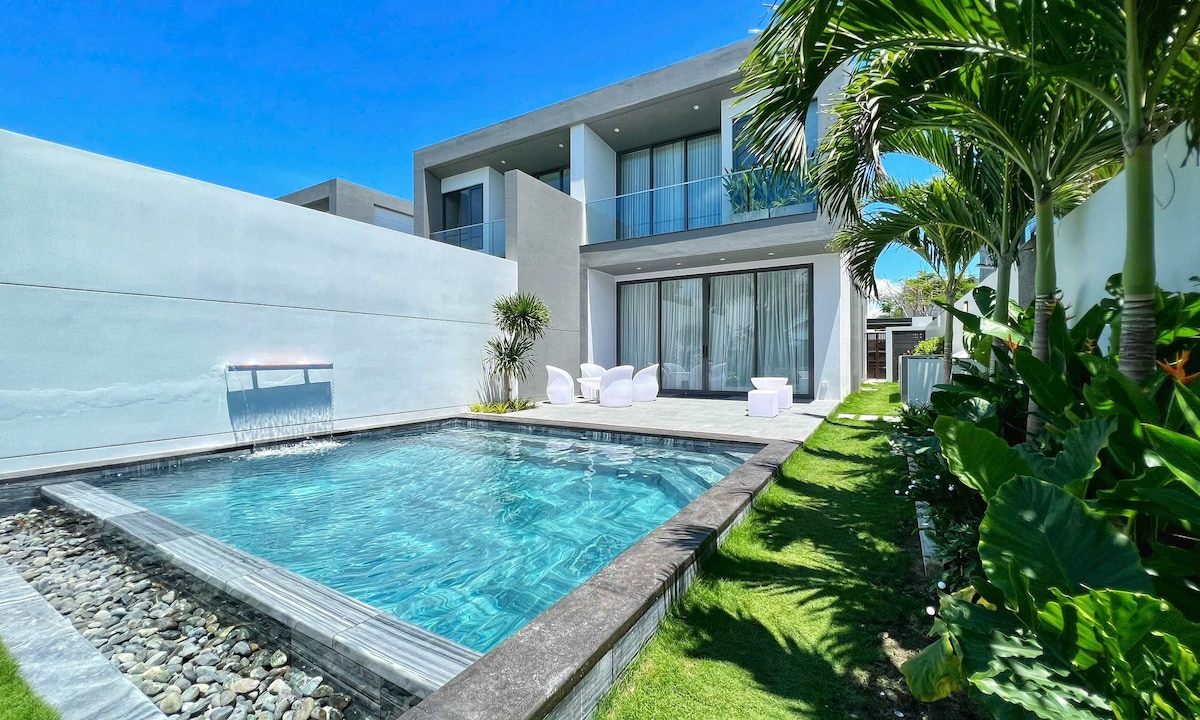 A modern villa features a private swimming pool surrounded by a well-kept garden. The exterior showcases large glass doors that lead to a spacious outdoor area, with comfortable white seating positioned nearby. Lush green plants frame the villa, enhancing the tranquil environment.
