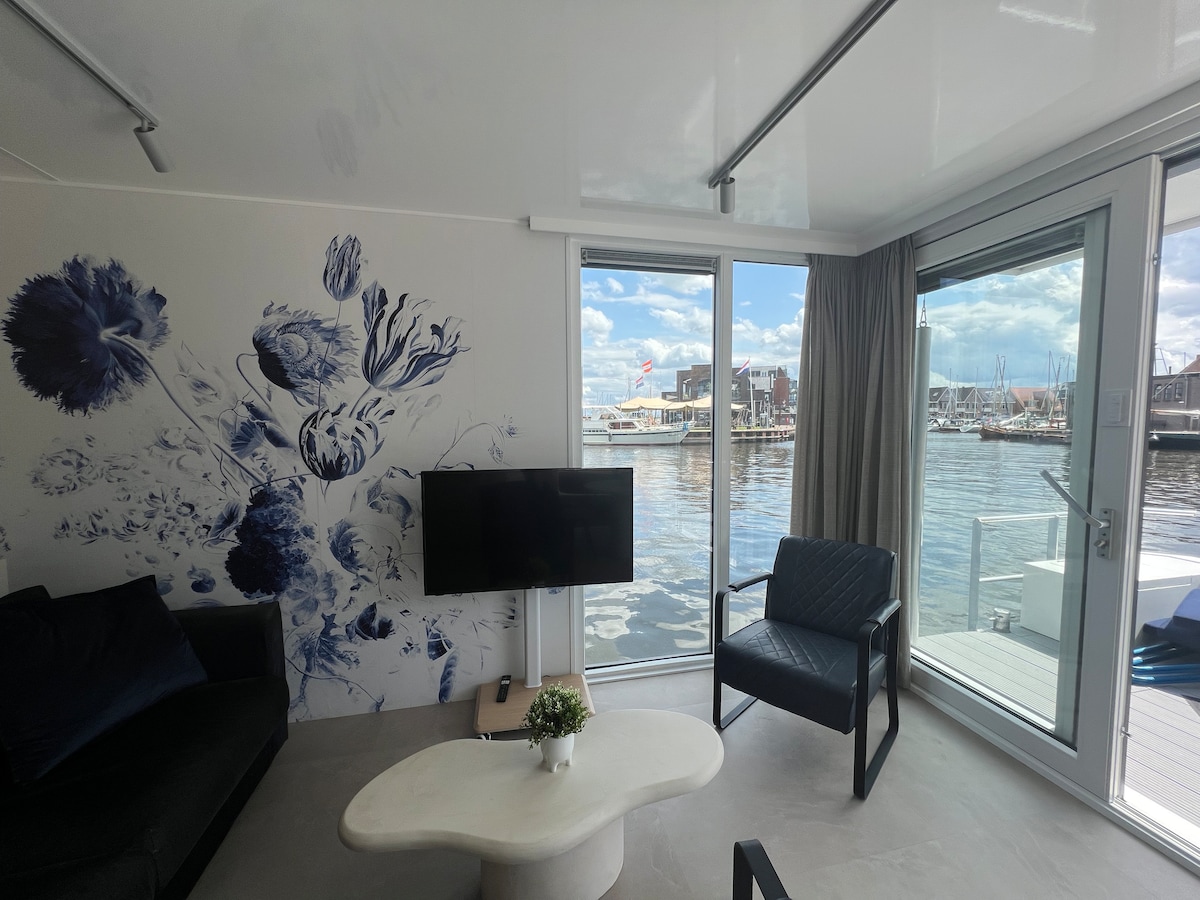 A modern living space features a large wall mural of blue flowers and a flat-screen television. Natural light floods in through large glass doors, which offer views of the harbor and boats, complemented by a cozy seating area that includes a black armchair and a coffee table.
