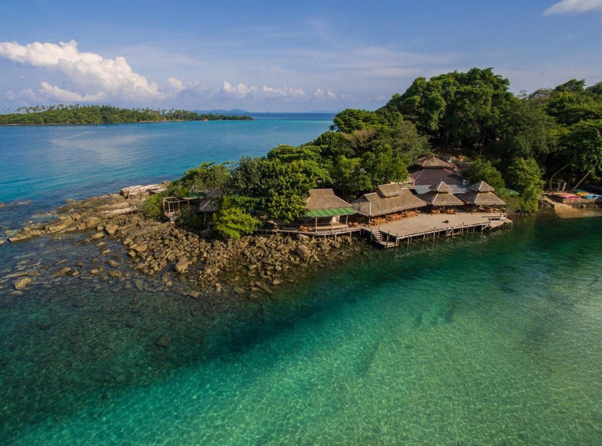An aerial view showcases a cluster of thatched-roof structures situated on a rocky shoreline, surrounded by turquoise waters. Lush greenery envelopes the area, blending harmoniously with the vibrant seascape under a clear sky dotted with clouds.