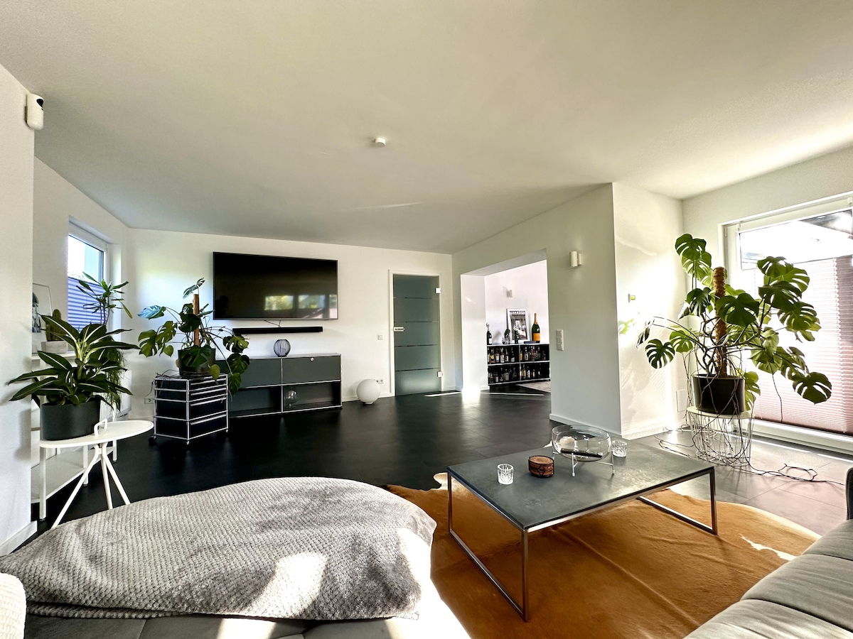 A spacious living area is shown, featuring a large wall-mounted TV and a sleek entertainment unit. Lush green plants are positioned throughout, adding a touch of nature. A coffee table rests on a stylish area rug, and large windows provide natural light while revealing sliding doors that lead to the terrace.