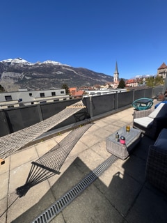 A spacious balcony features a lounge area with modern wicker furniture and a low table, set against a backdrop of mountain views. A hammock is positioned nearby, along with two bottles positioned on the table, inviting relaxation under clear blue skies.