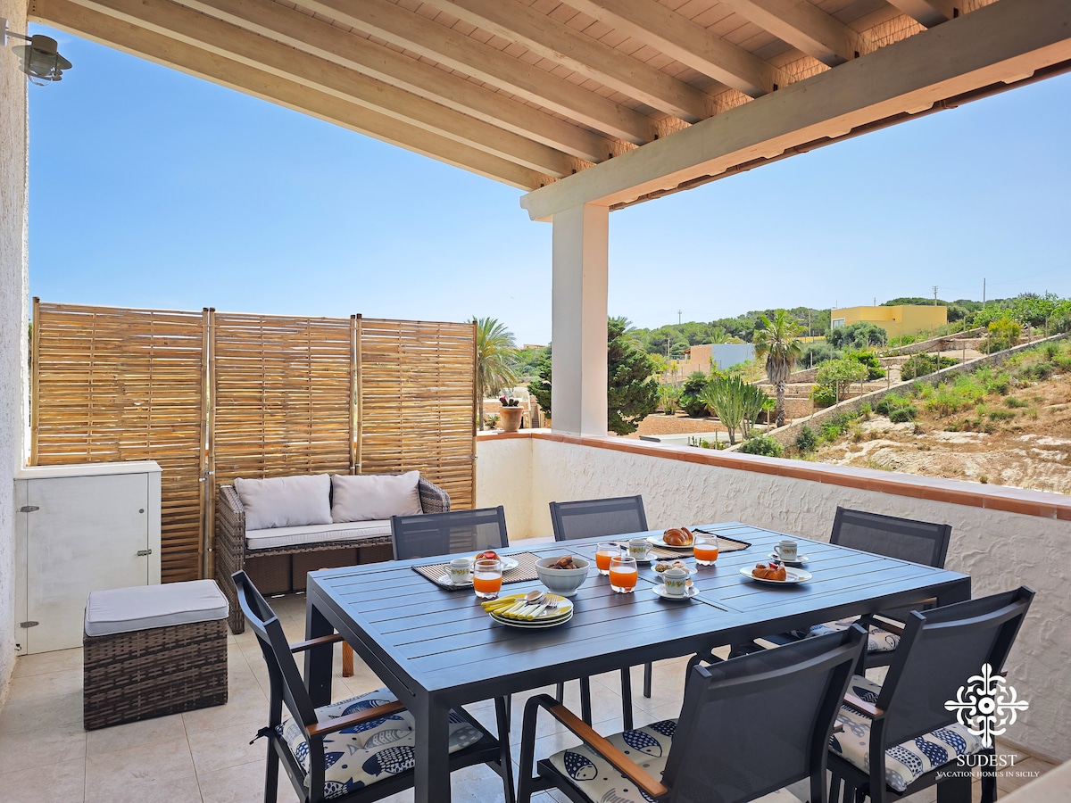 A covered veranda is furnished with a spacious dining table surrounded by several chairs, ideal for outdoor meals. A sitting area with a cushioned sofa offers a relaxed space to enjoy the surrounding greenery and views of the landscape.