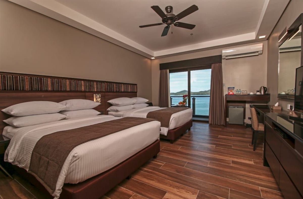 A spacious room features two queen-sized beds with crisp white linens, situated on a wood-floored surface. Large windows offer views of the bay, while a mounted flat-screen television and a work desk are positioned against the wall. A small refrigerator and coffee-making facilities are also included.