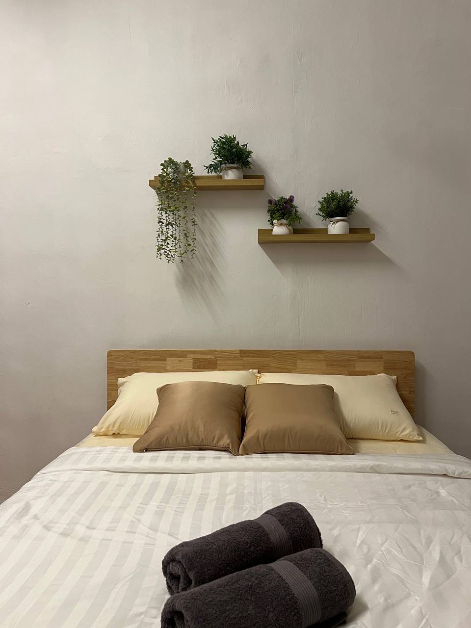 A well-lit sleeping area features a wooden headboard with a spacious bed dressed in white linens. Two decorative shelves mounted on the wall display small potted plants, adding a touch of greenery. Soft towels are neatly rolled at the foot of the bed.