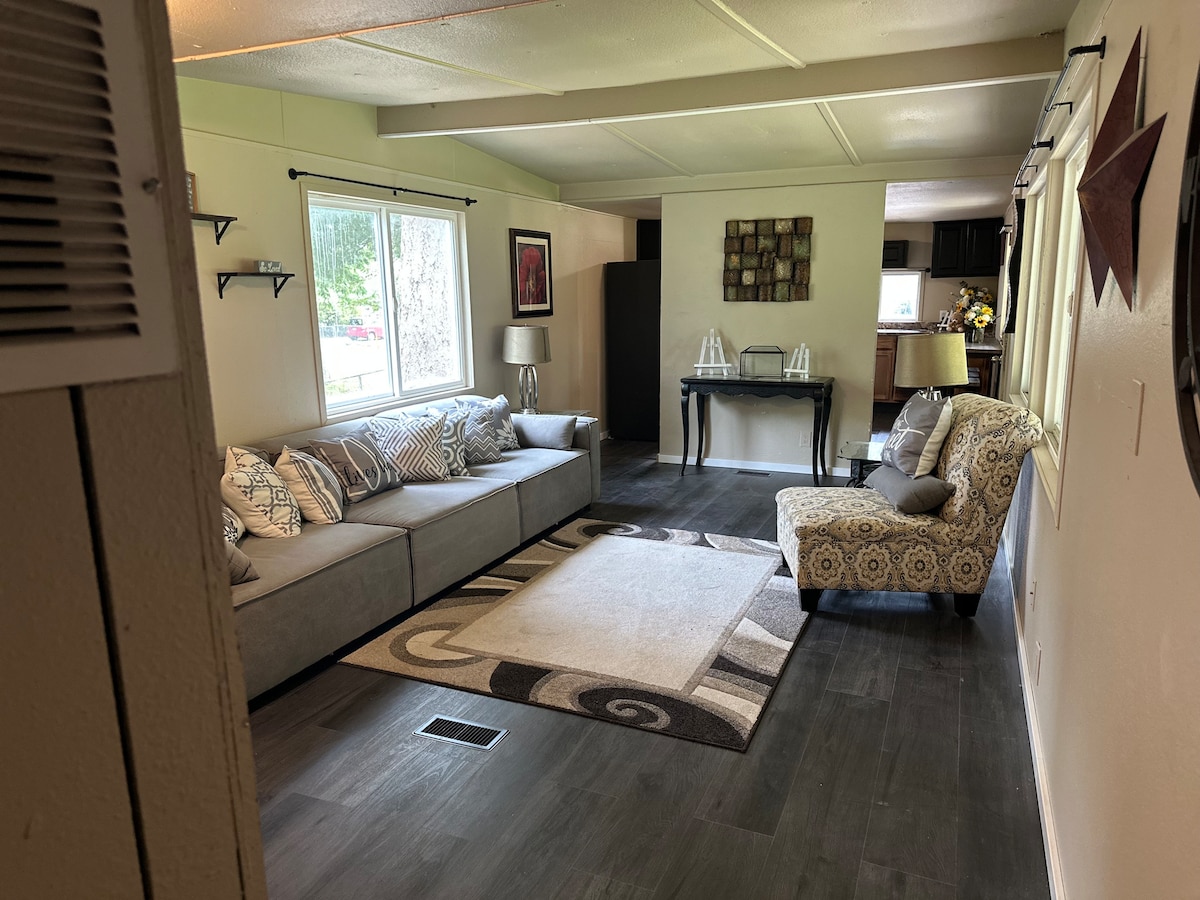 A spacious living area is highlighted by a large sectional sofa adorned with patterned cushions. Light enters through a large window, illuminating a central area rug and a cozy accent chair. A dining space can be seen in the background, featuring a dark table and chairs.