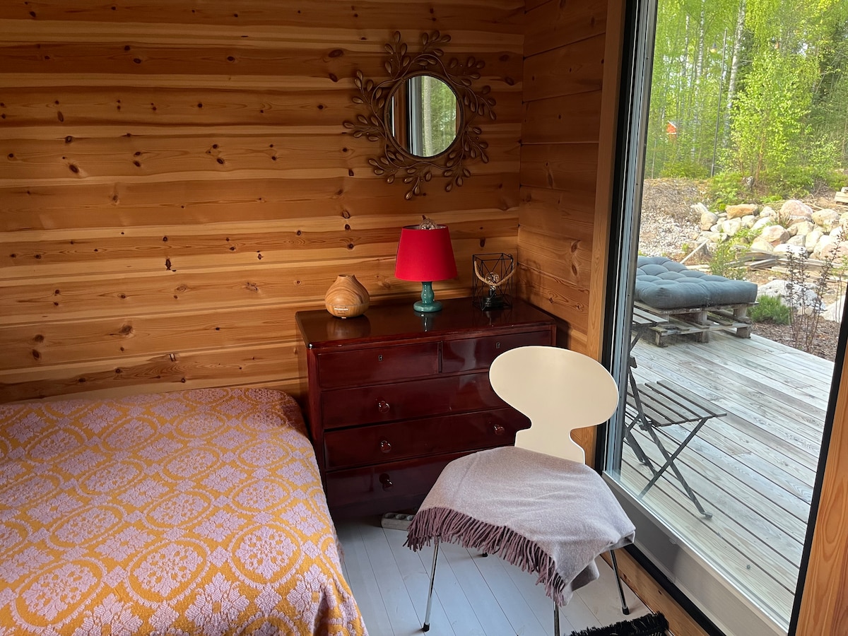 A warm wooden interior features a spacious bed adorned with a patterned blanket and a bedside table with a lamp. A chair and a small throw blanket are positioned beside the bed. Floor-to-ceiling windows reveal access to an outdoor deck surrounded by trees.