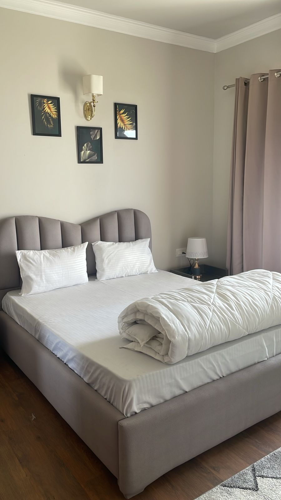 A cozy bedroom is presented with a comfortable bed featuring a plush headboard. Two decorative pillows adorn the bed, and a neatly rolled blanket is placed on top. Soft lighting is provided by a bedside lamp, while framed artwork adds a touch of elegance to the walls.