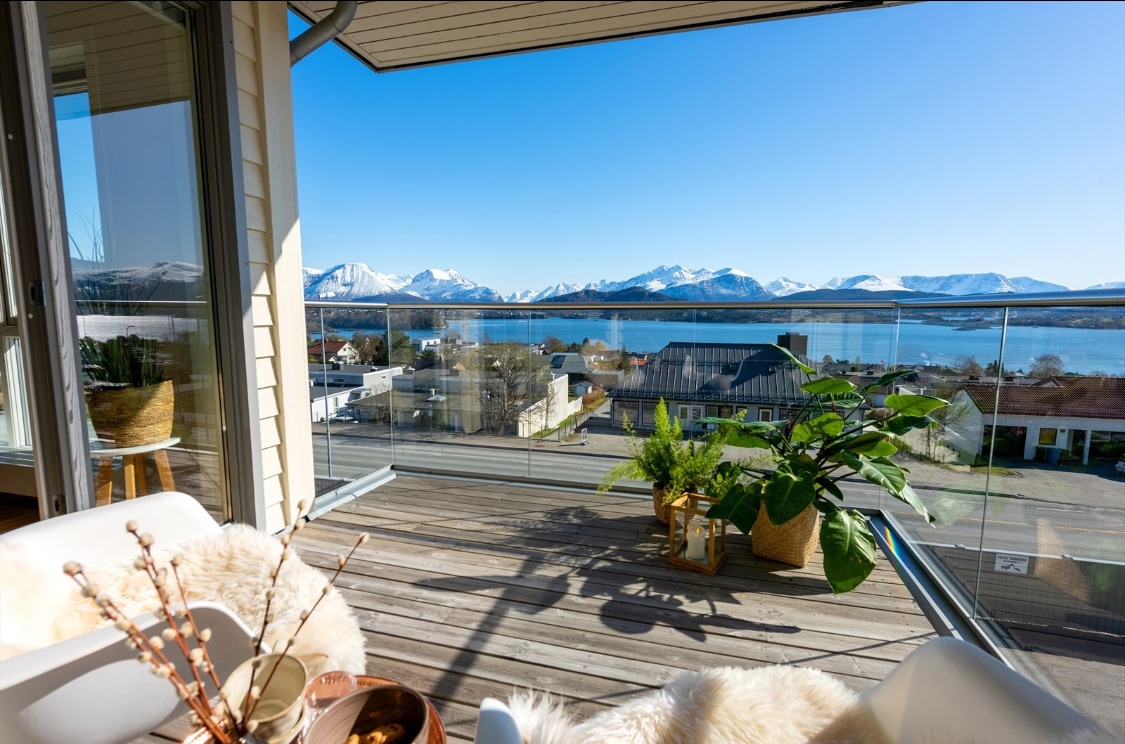 A balcony is displayed with glass railings, offering unobstructed views of distant mountains and a serene fjord. Potted plants are positioned on the wooden floor, and a comfortable seating area is arranged, providing an inviting space to enjoy the scenery.