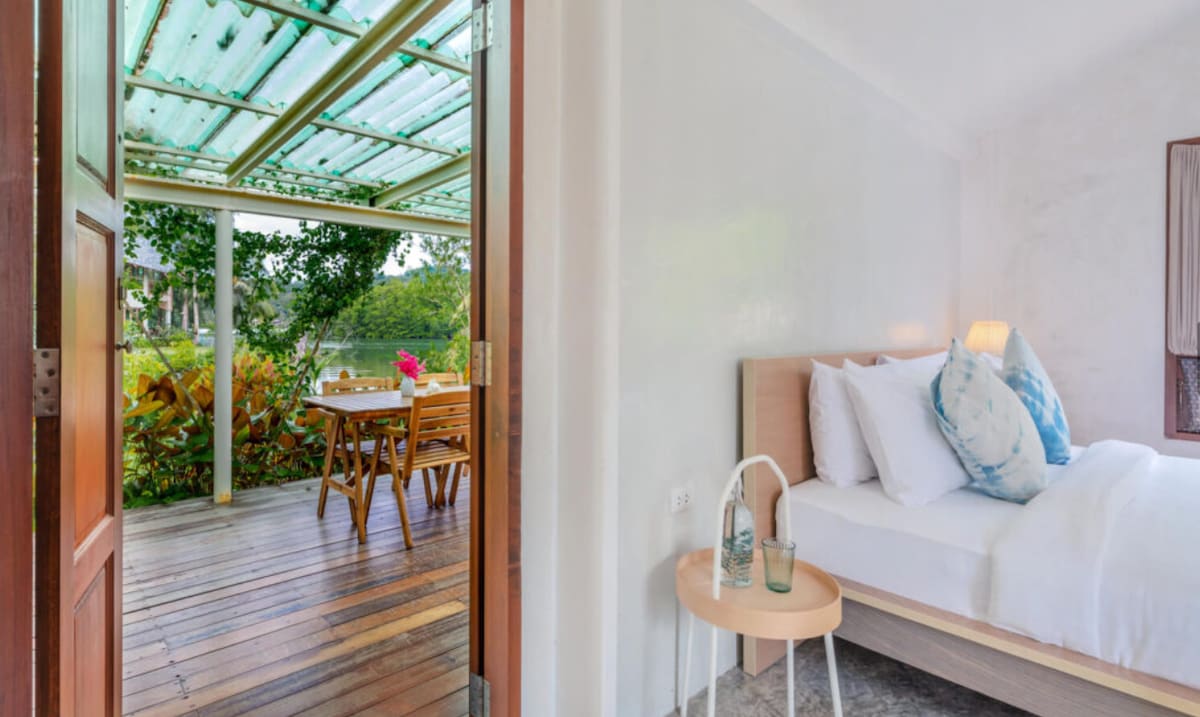 A doorway connects a cozy sleeping area with a private outdoor space. The room features a light-colored bed adorned with patterned pillows. A wooden table and chairs are visible on the shaded deck, surrounded by lush greenery, inviting relaxation.