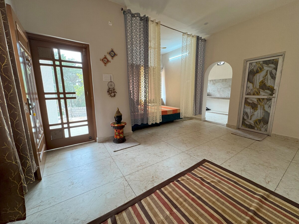 The bright studio space features a large entrance door and large windows, allowing natural light to fill the room. A colorful decorative piece stands next to the door, while a striped rug adds comfort to the tiled floor. Dividers create distinct areas within the room.