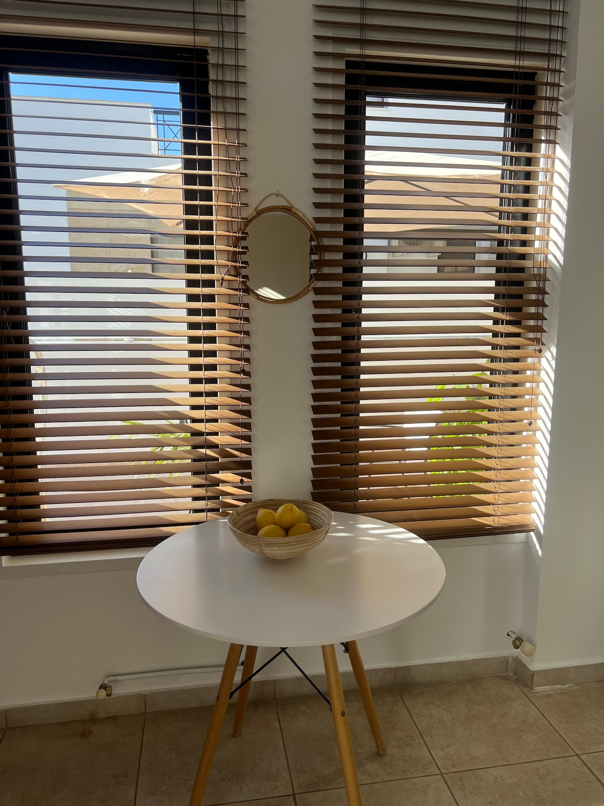 A small round table is positioned beneath two windowed openings, outfitted with horizontal wooden blinds. A simple mirror hangs on the wall, reflecting light. A basket containing lemons is placed on the table, complementing the bright and airy ambiance.