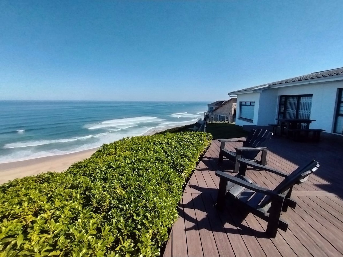 A deck area is seen overlooking the ocean, featuring two black lounge chairs positioned for optimal sea views. Lush green foliage frames the space, and gentle waves can be observed on the horizon under a clear blue sky.