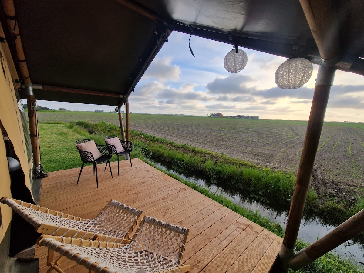 A spacious wooden terrace outside a safari tent features two black wire chairs and a natural woven lounge chair. The surrounding landscape showcases open fields under a cloudy sky, with subtle hues of green and brown visible in the distance.