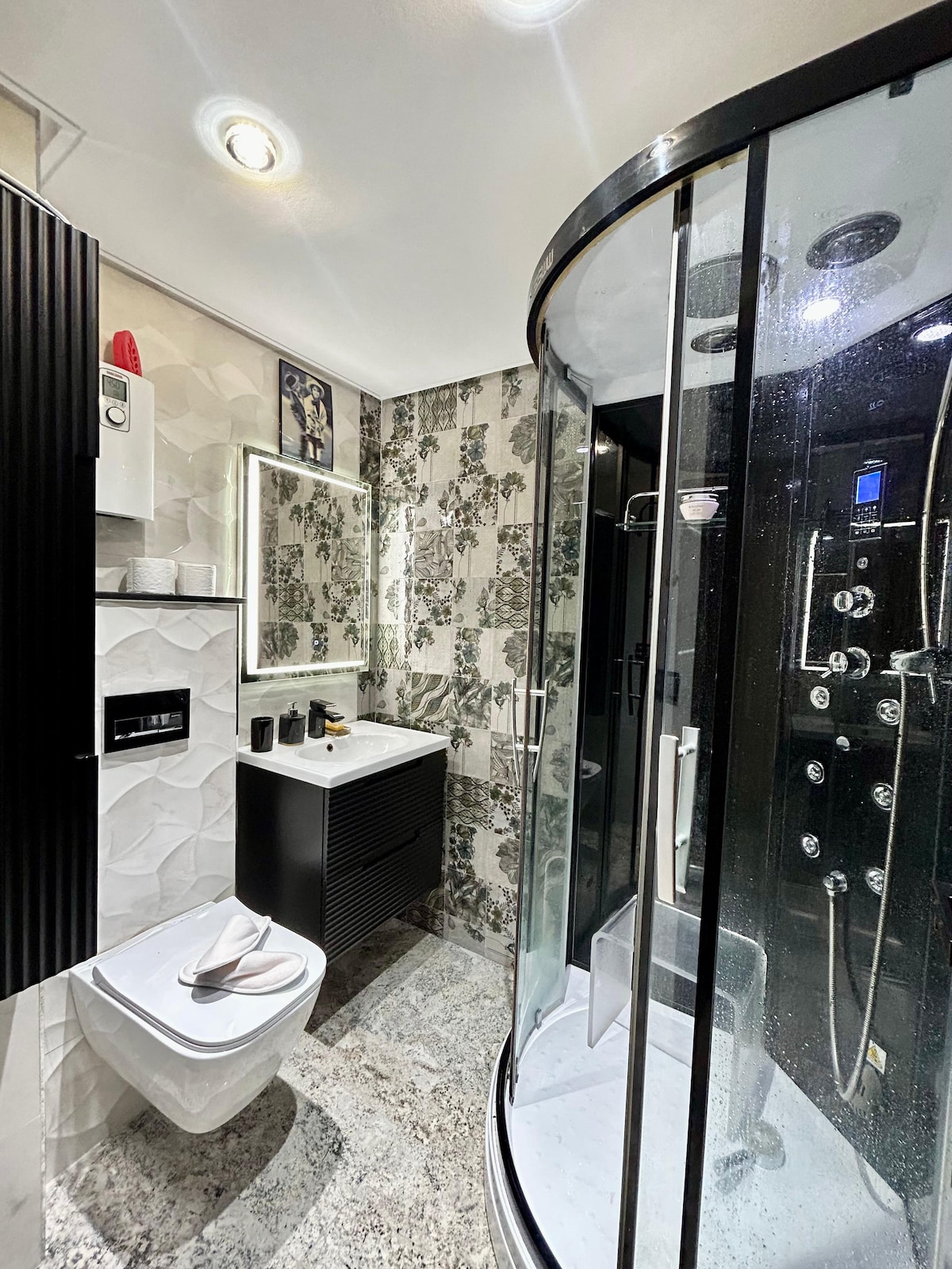 A modern bathroom features a sleek shower enclosure with multiple shower heads. A compact vanity with a large mirror is positioned beside a stylish toilet. Elegant black and white tiles create a refined ambiance, while natural light brightens the space.