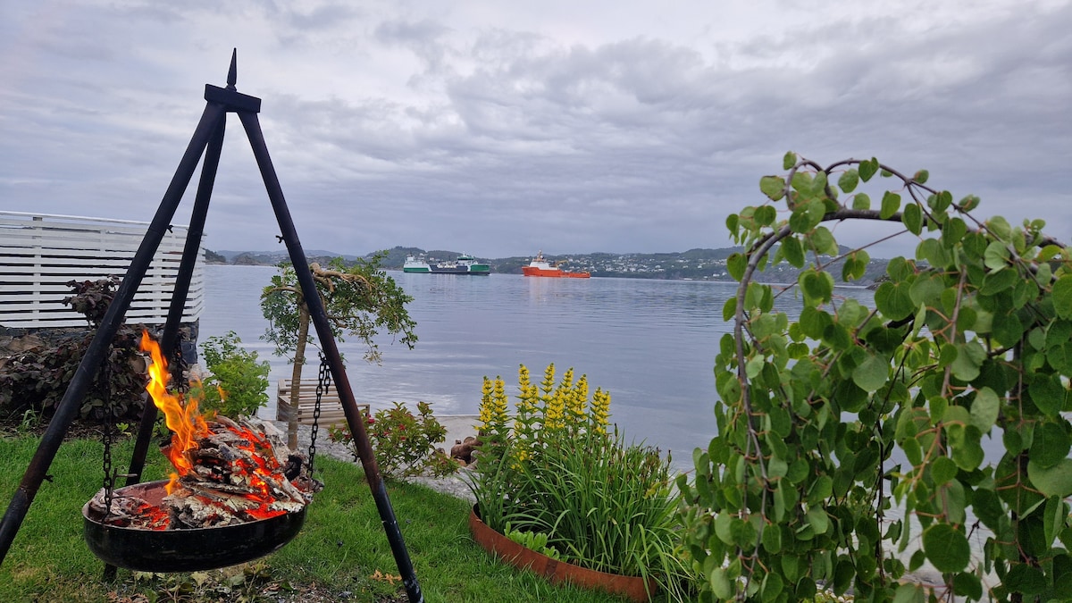 A fire pit is seen in a scenic outdoor space, surrounded by greenery and flowers. Calm waters reflect a cloudy sky, with distant boats visible on the horizon. The area is designed for relaxation and enjoyment by the water.