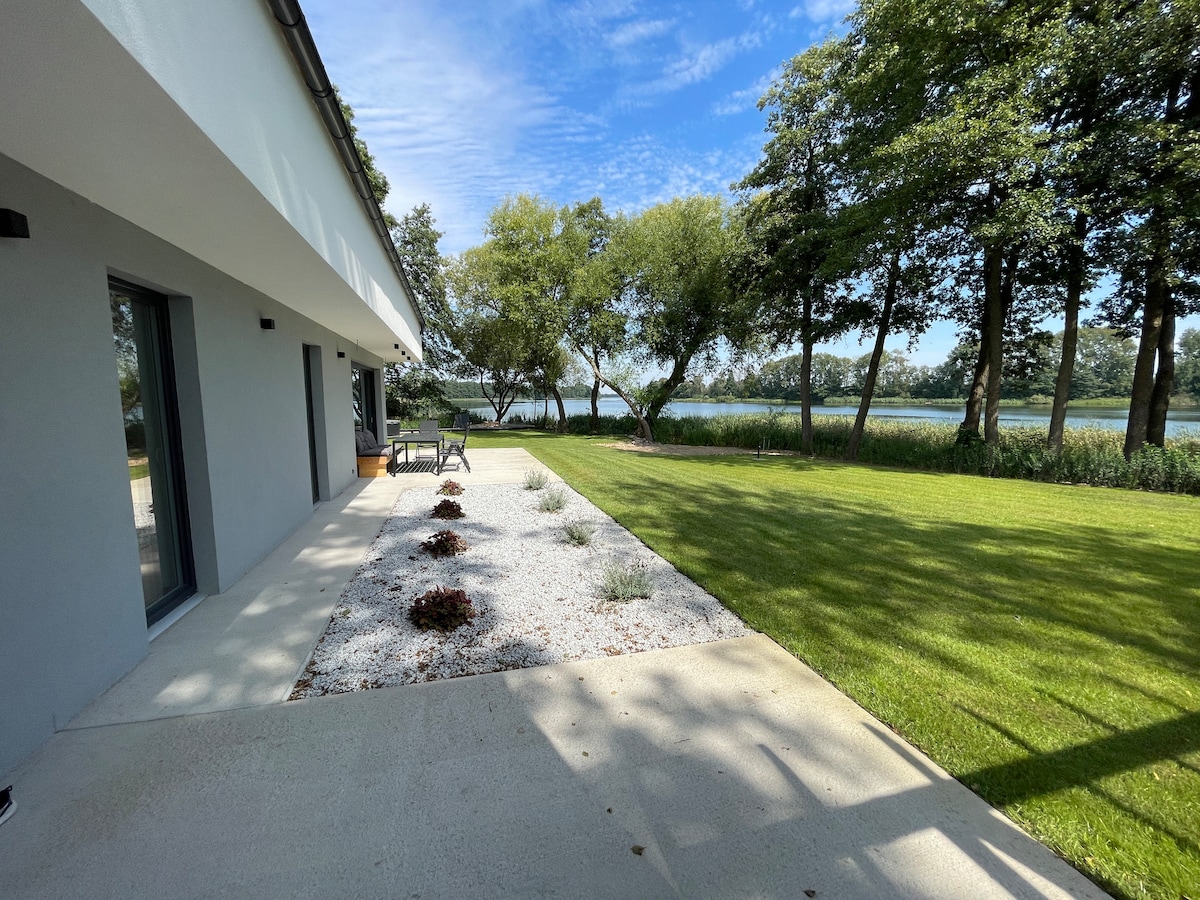 A serene outdoor space showcases a well-maintained lawn bordered by decorative gravel and small plantings. Sunlight filters through the trees, creating a calm atmosphere. The patio area features seating options, providing a peaceful spot to enjoy the scenic views of the nearby lake.