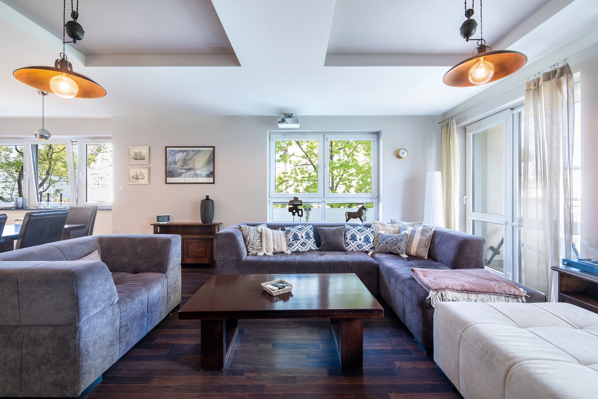 A spacious living area features a large sectional sofa adorned with decorative pillows, accompanied by a wooden coffee table. Natural light floods the room through multiple windows, which offer views of greenery. Subtle pendant lights hang from the ceiling, creating a comfortable ambiance.