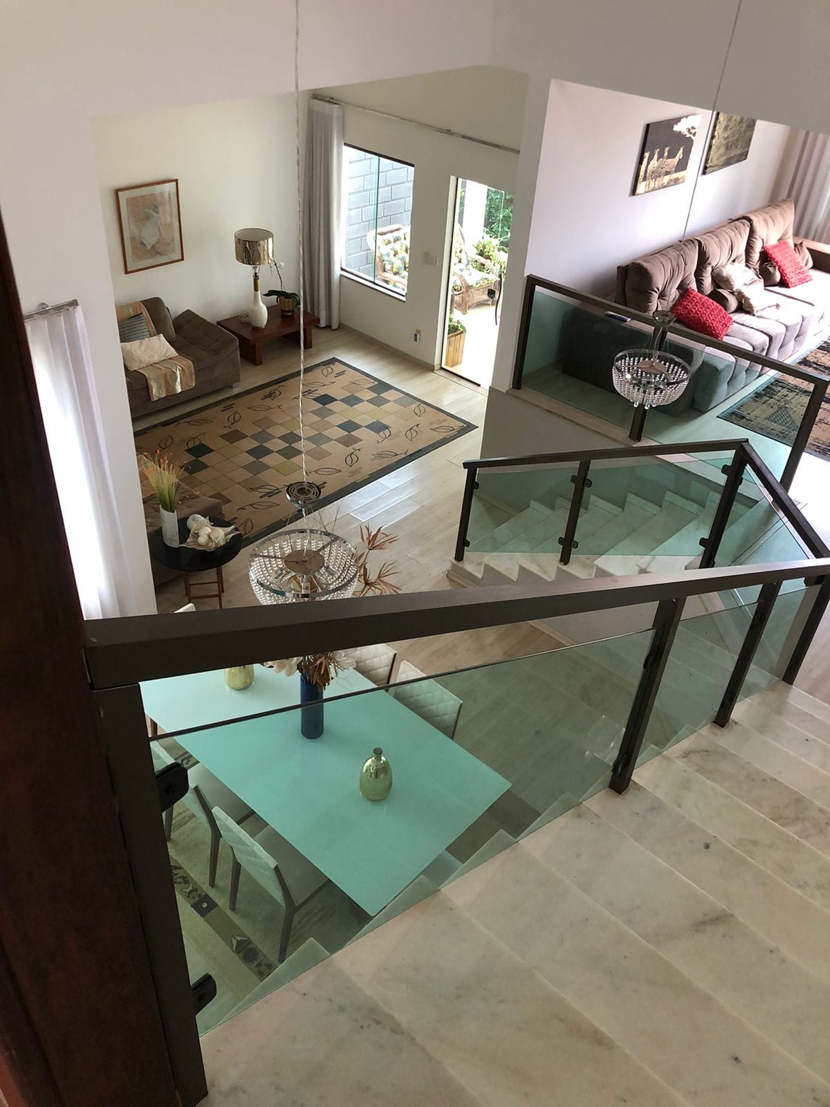 A multi-level interior view showcases an open layout, with a glass railing staircase leading to a dining area below. Natural light fills the space, highlighting a light blue table set beneath a decorative vase. Cozy seating areas are visible alongside a patterned rug.