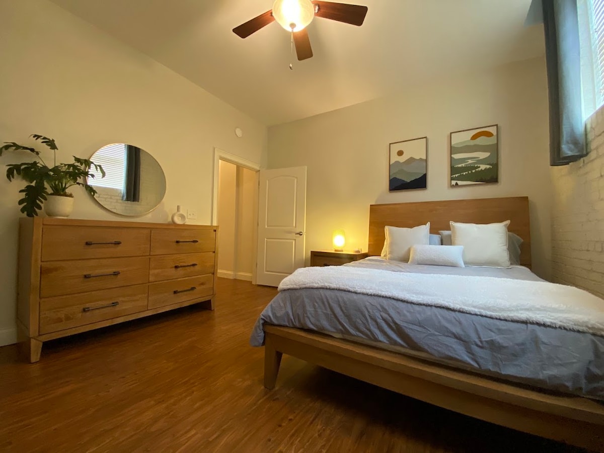 A serene bedroom features a spacious wooden bed with white pillows and a soft throw. A light wood dresser and a round mirror are positioned against a light-colored wall. Two framed artworks are displayed above the bed, complemented by natural light from a window.