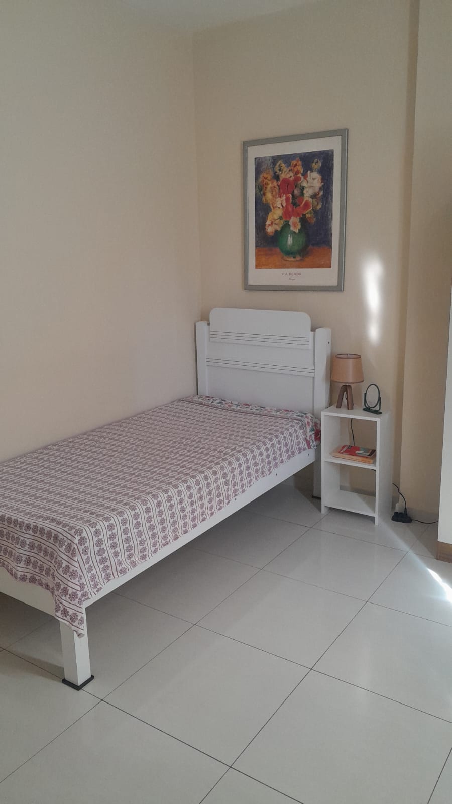 A bright sleeping area features a single bed draped with a patterned blanket. A bedside table holds a lamp, and a piece of vibrant artwork adorns the wall above. The floor is tiled, reflecting natural light streaming in from a nearby window.