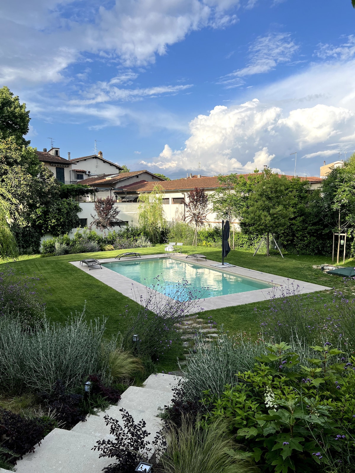 A tranquil outdoor area is depicted, featuring a clear swimming pool surrounded by lush greenery and well-maintained grass. Various trees and plants frame the scene, while patio furniture is placed nearby, inviting relaxation. A swing set adds a playful touch to the space.