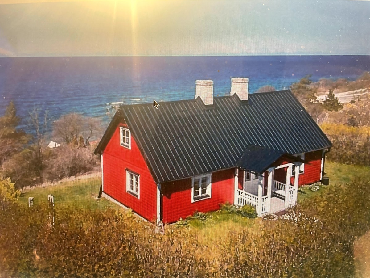 A charming red house is situated on a hillside overlooking the ocean, framed by green foliage. The structure features a black roof and white accents, including a small porch. Natural light from the sun casts a warm glow over the landscape.