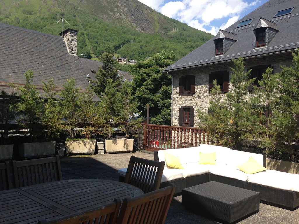 An expansive terrace is shown, featuring a comfortable seating area with light-colored cushions and a dark coffee table. Surrounding greenery and stone buildings are visible, set against a backdrop of a mountainous landscape under a clear blue sky.