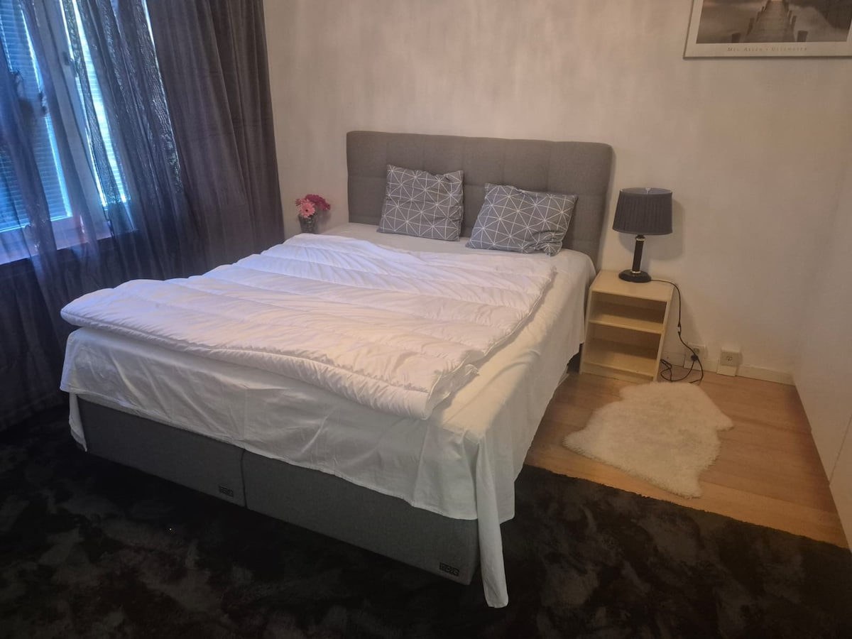 A cozy sleeping area features a spacious bed with a white comforter, complemented by decorative pillows. A bedside table with a lamp sits next to the bed. Natural light filters in through the window, covered by sheer curtains, casting a soft glow in the room.