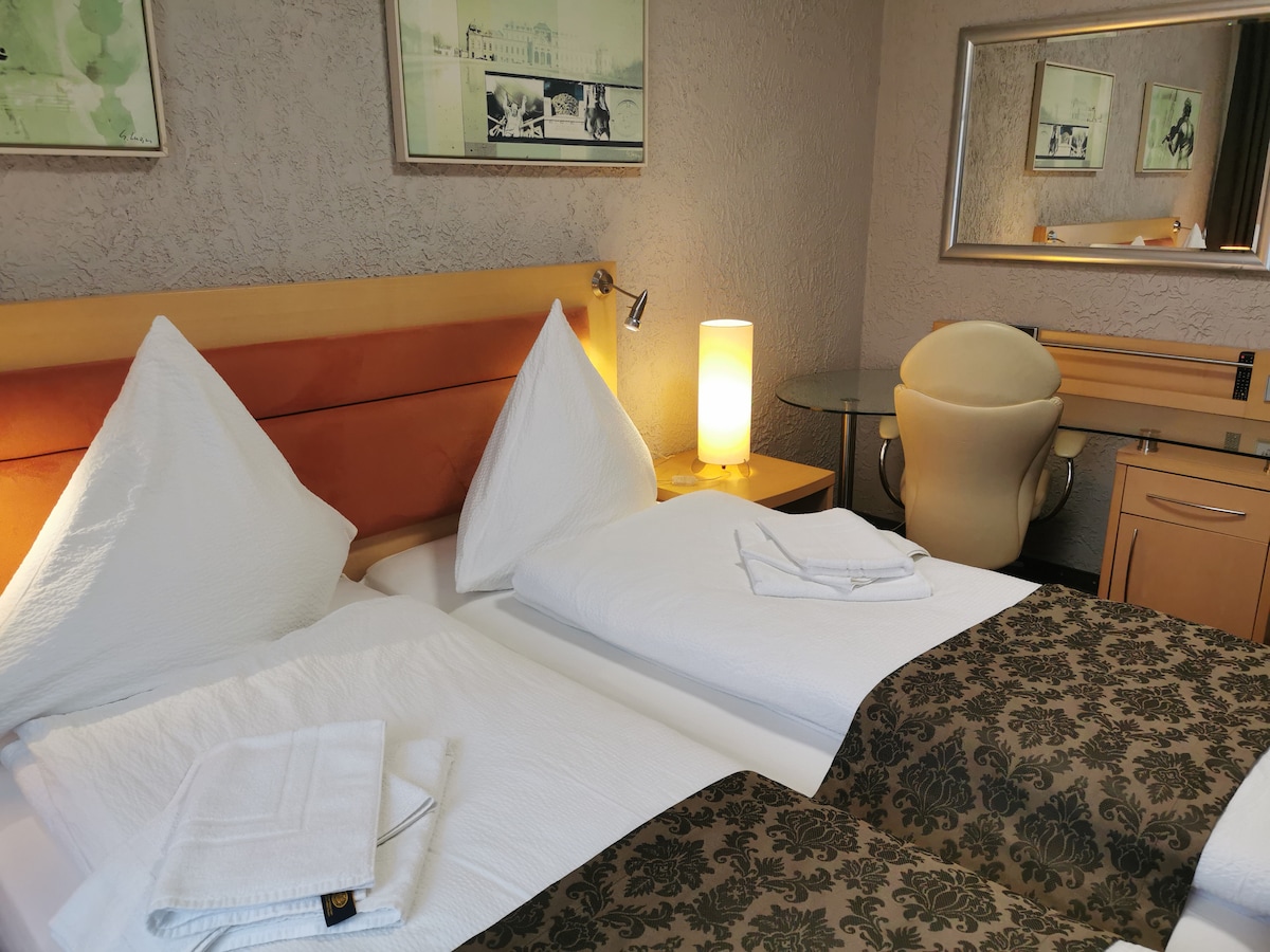 The sleeping area features two spacious beds with white linens and decorative pillows. A bedside lamp provides soft lighting. A mirrored wall reflects a desk and chair, enhancing the room's functionality. Artwork is displayed above the beds, adding a decorative touch to the space.