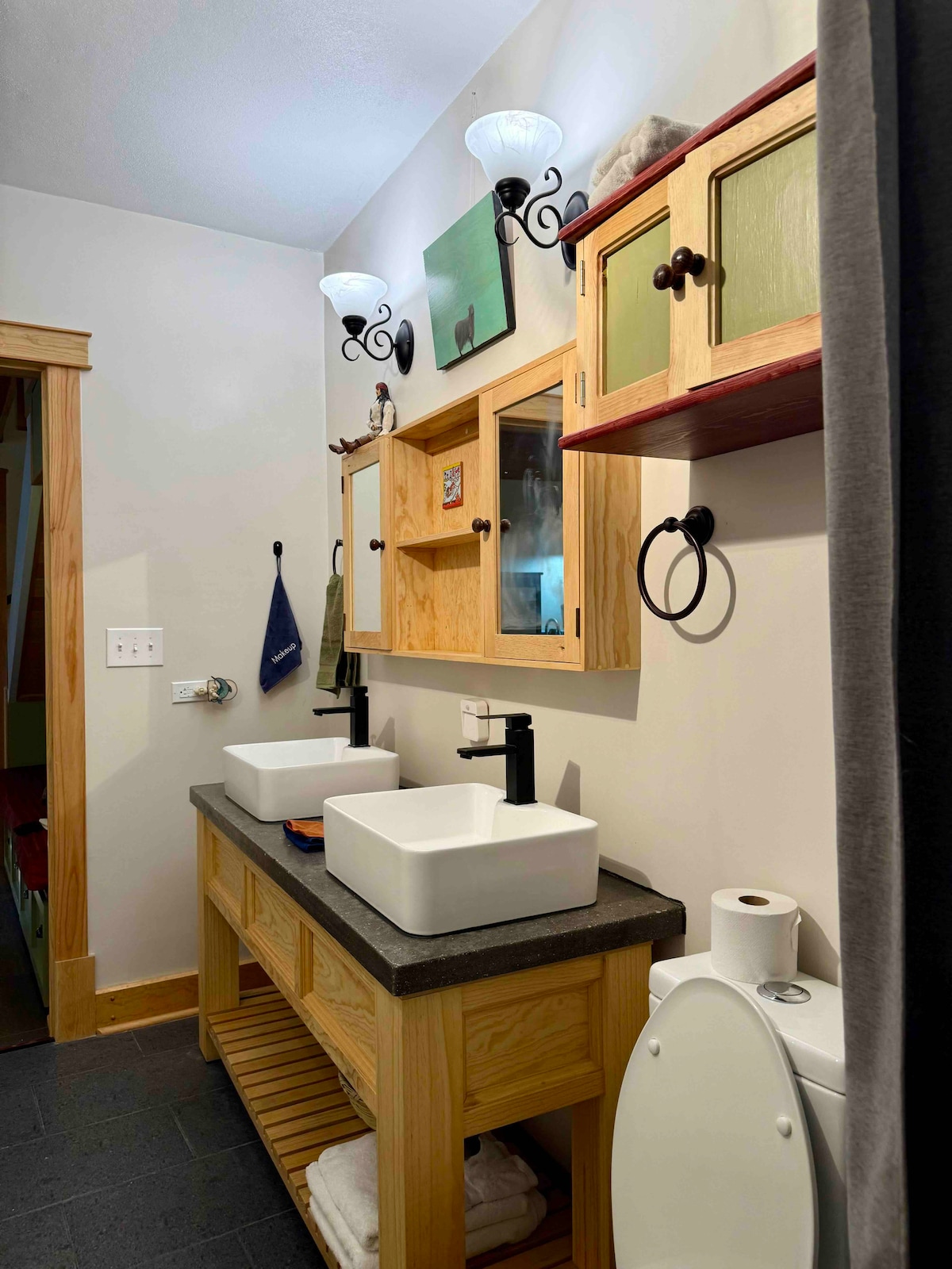 A well-organized bathroom is depicted, featuring a dual sink setup with modern fixtures. Above the sinks, cabinetry provides storage space. Wall-mounted sconces offer illumination. A roll of toilet paper and a stack of towels are visible, contributing to the practicality of the space.