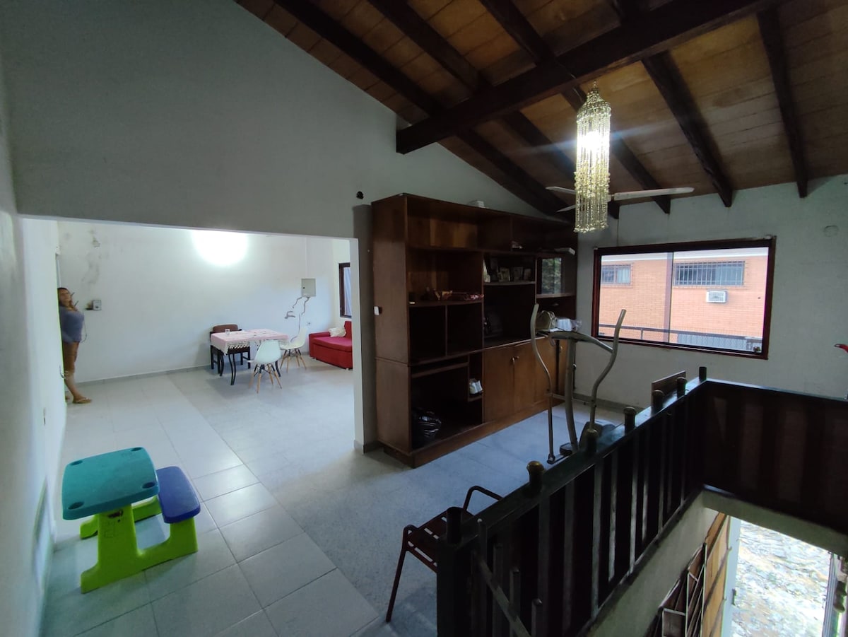 An open living space is viewed from an elevated angle, showcasing a combination of the dining area and a cozy seating arrangement. A wooden bookshelf with decorative items lines one wall, while a small table for two is positioned between the areas. Natural light illuminates the space through large windows.