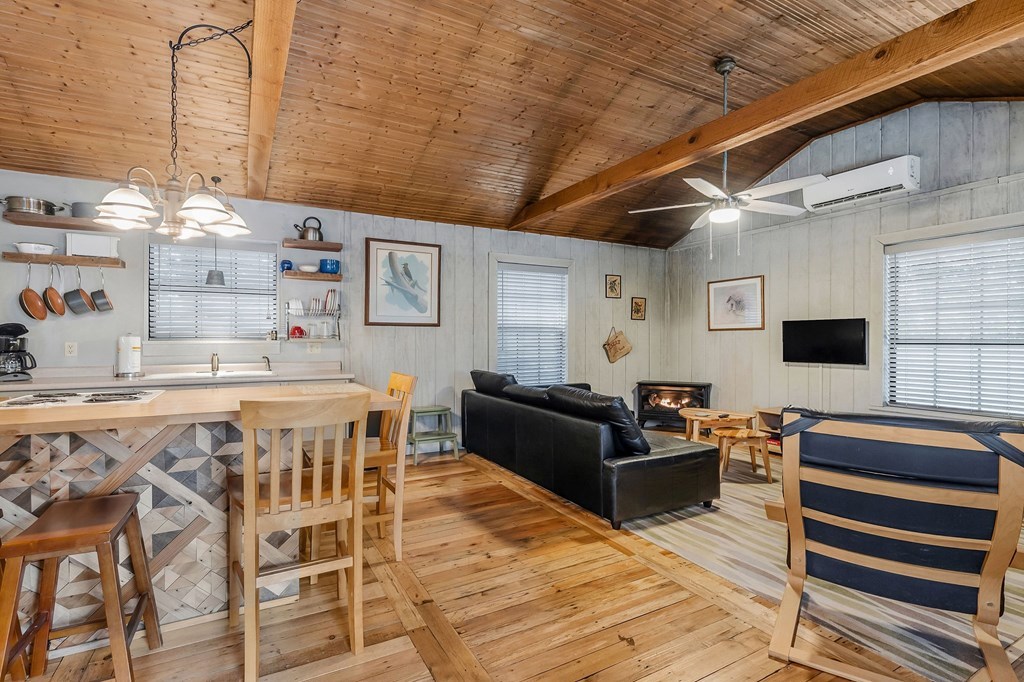 The open living area is characterized by a wood-paneled ceiling with exposed beams. A comfortable black sofa and a wooden table with chairs are visible. A flat-screen TV is mounted on the wall, while a small electric fireplace adds a cozy touch to the space.
