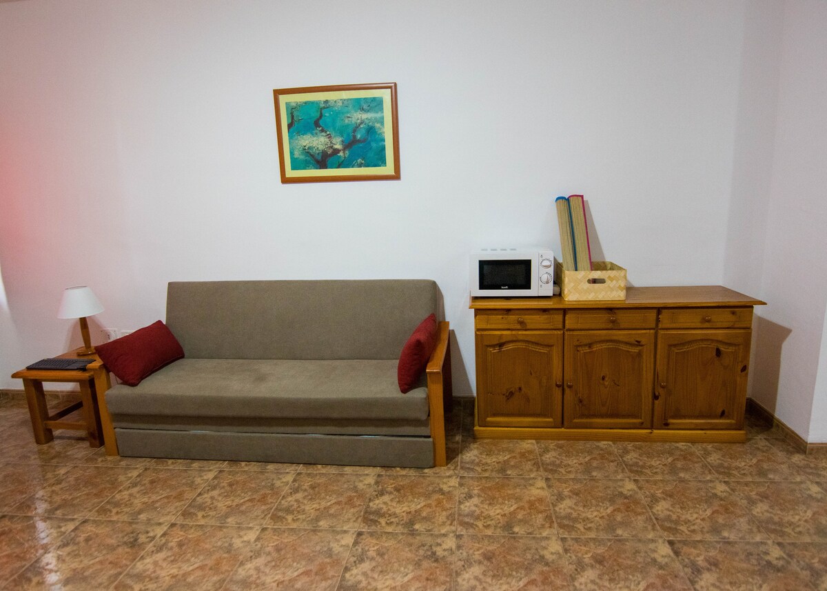 A living area features a comfortable sofa with red accent pillows and a side table. A wooden cabinet is against the wall, accompanied by a microwave and a decorative basket. An artwork hangs above the sofa, adding a touch of color to the neutral space.
