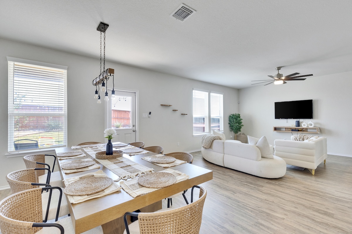 An open-concept living area is showcased, featuring a spacious dining table with multiple woven chairs and a cozy sectional sofa. Natural light fills the room through large windows, which frame a view of the backyard. A ceiling fan circulates air while a wall-mounted TV adds convenience.