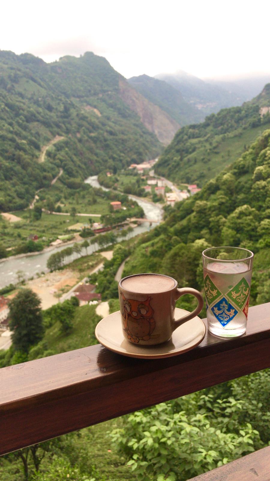 A scenic view of a river winding through lush green valleys is visible. A mug and a glass are placed on a wooden surface in the foreground, complementing the tranquil landscape. The misty mountains rise in the background, adding depth to the scene.