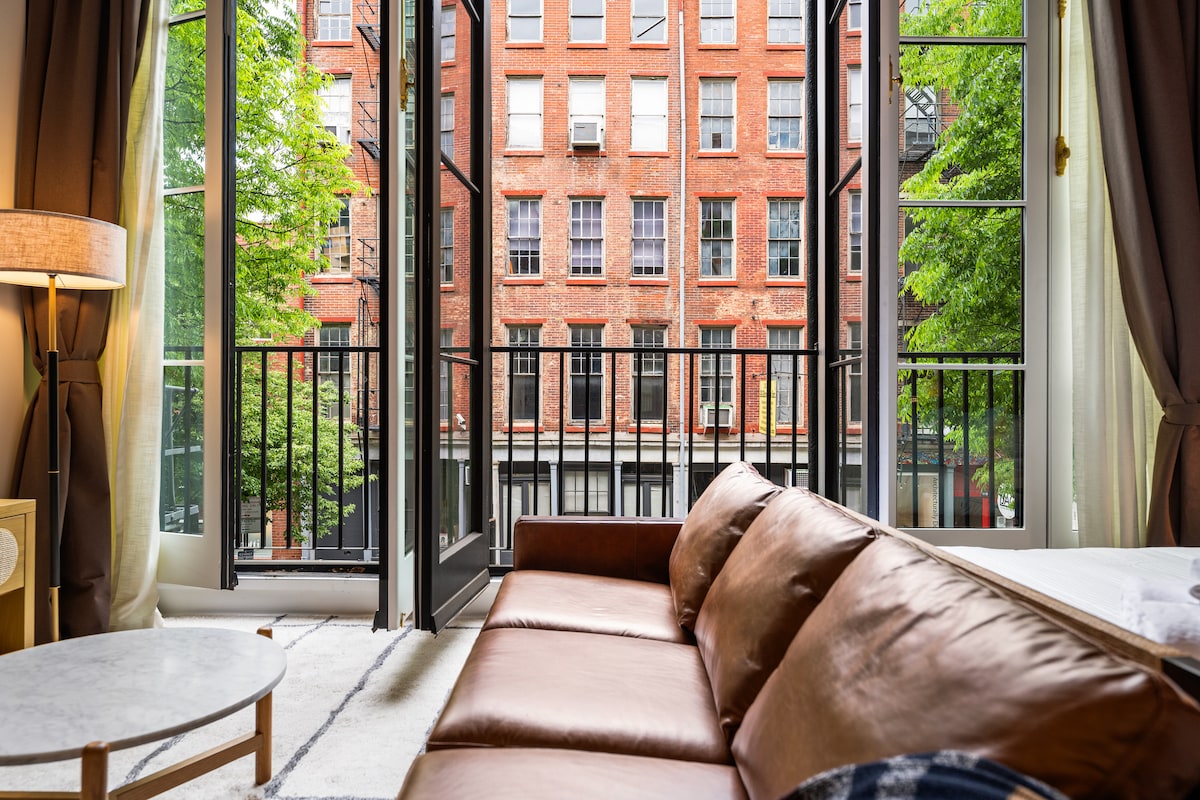 Large windows frame a view of a historic brick building across the street, allowing natural light to fill the room. A brown leather sofa is positioned in front of the windows, with a round marble coffee table nearby. Greenery from trees outside adds a refreshing touch.