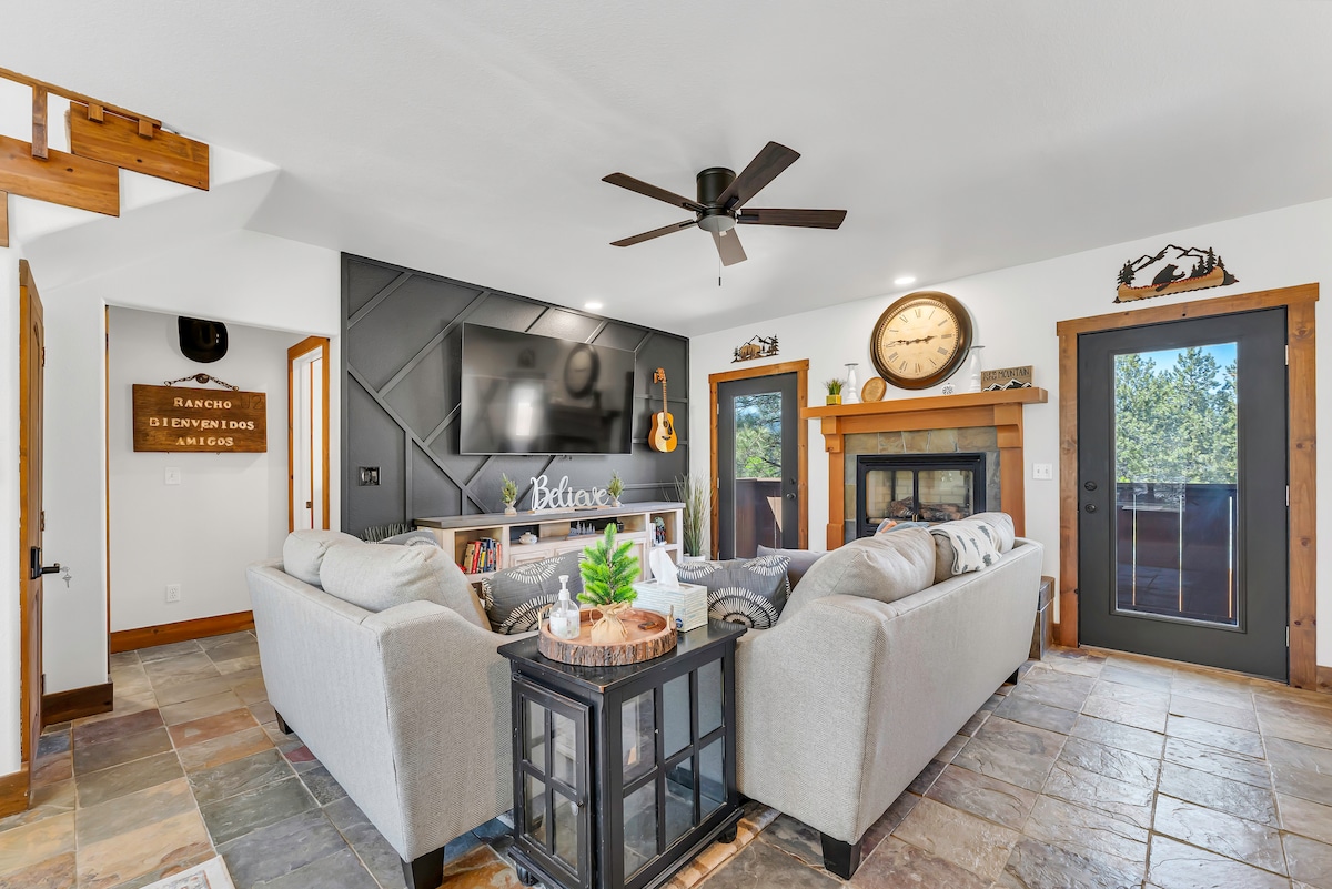 A comfortable living area is highlighted by a gray sectional sofa, facing a wall-mounted television and a cozy fireplace. A large decorative clock and a coffee table with plants add character to the space, complemented by natural light from the nearby door leading to the outdoors.