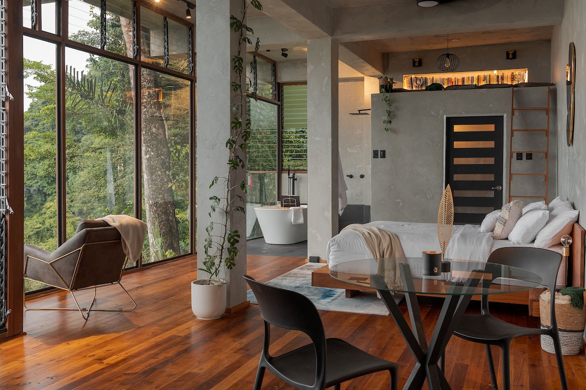 An open floor plan reveals a spacious bedroom with a large bed and a cozy armchair, surrounded by expansive windows that provide views of the lush rainforest. A modern dining table is situated nearby, while natural light filters through the space, highlighting the wooden floors.