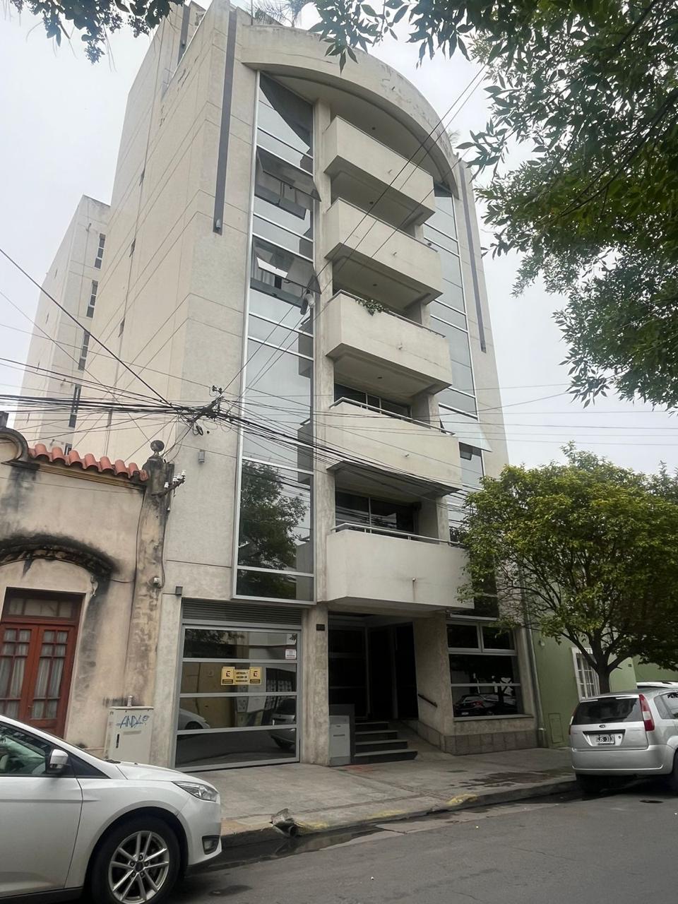 The exterior of a modern five-story building is presented, characterized by large glass windows and an arching balcony on the upper level. The entrance features a wide stairway, and surrounding greenery is visible, creating a blend of urban and natural elements.