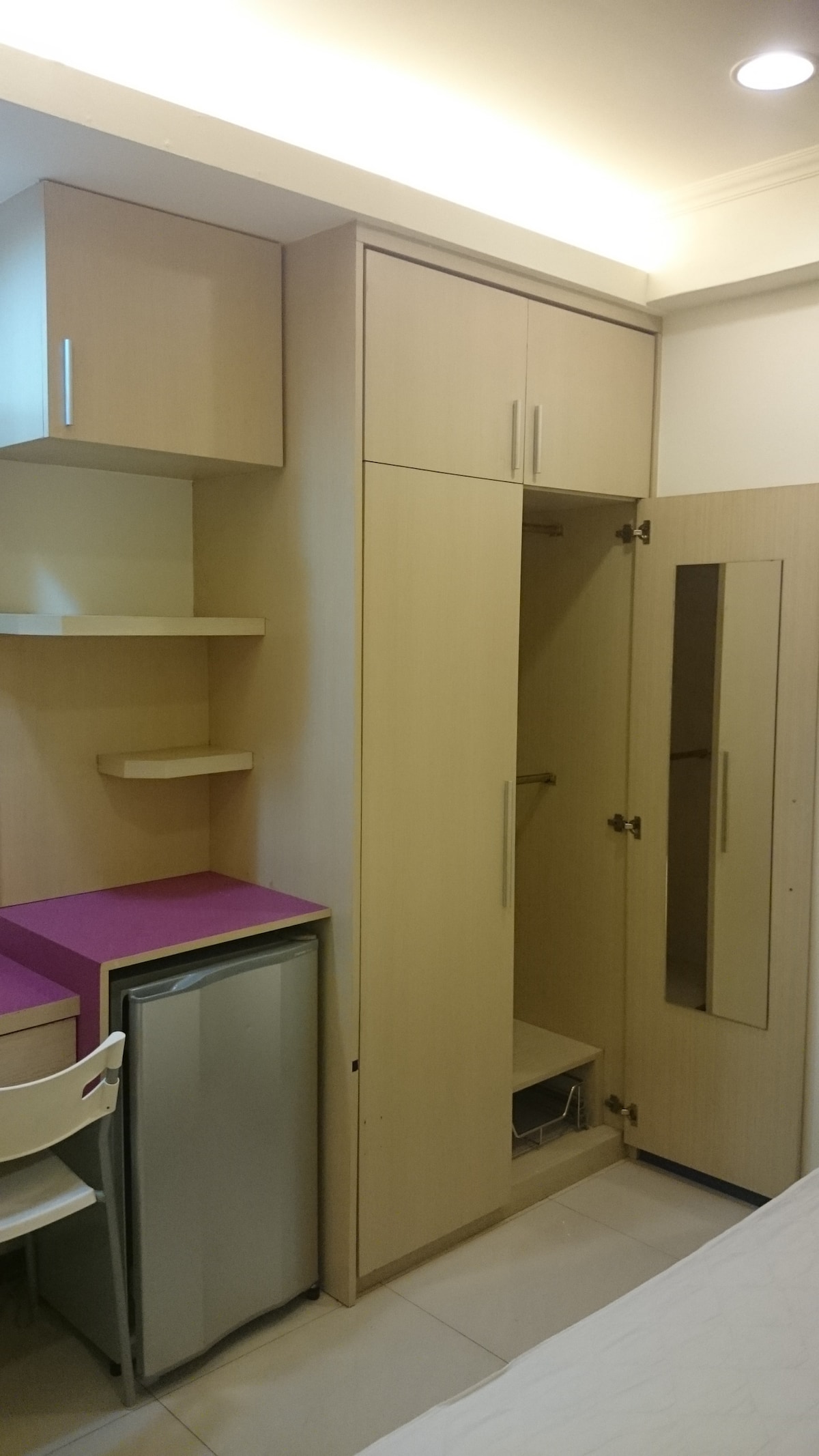 A compact living space features light wood cabinetry and a built-in closet. A small desk with a purple top is combined with a white chair, while a mini refrigerator is positioned nearby. Soft lighting enhances the functional layout of the area.