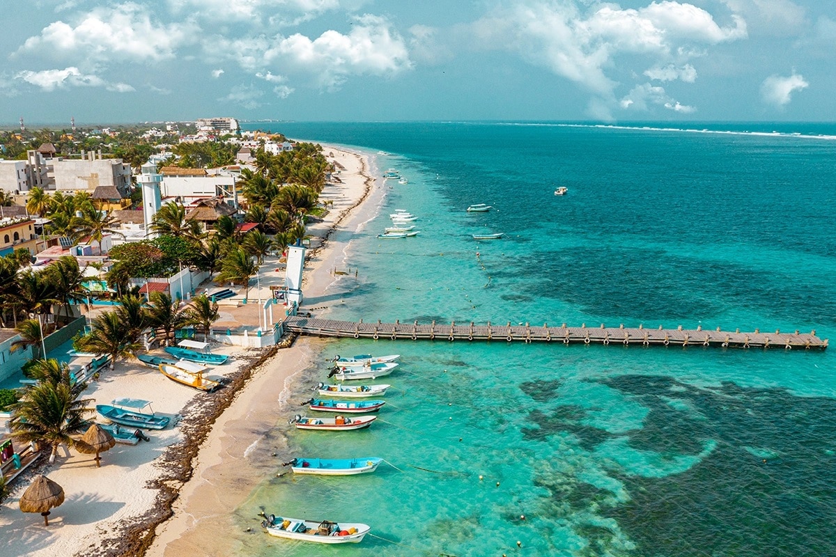 A scenic coastal view captures turquoise waters gently lapping against a sandy shore. Fishing boats are anchored near a wooden pier, with palm trees lining the beach. The landscape is framed by distant islands and cloudy skies, offering a serene atmosphere for visitors.