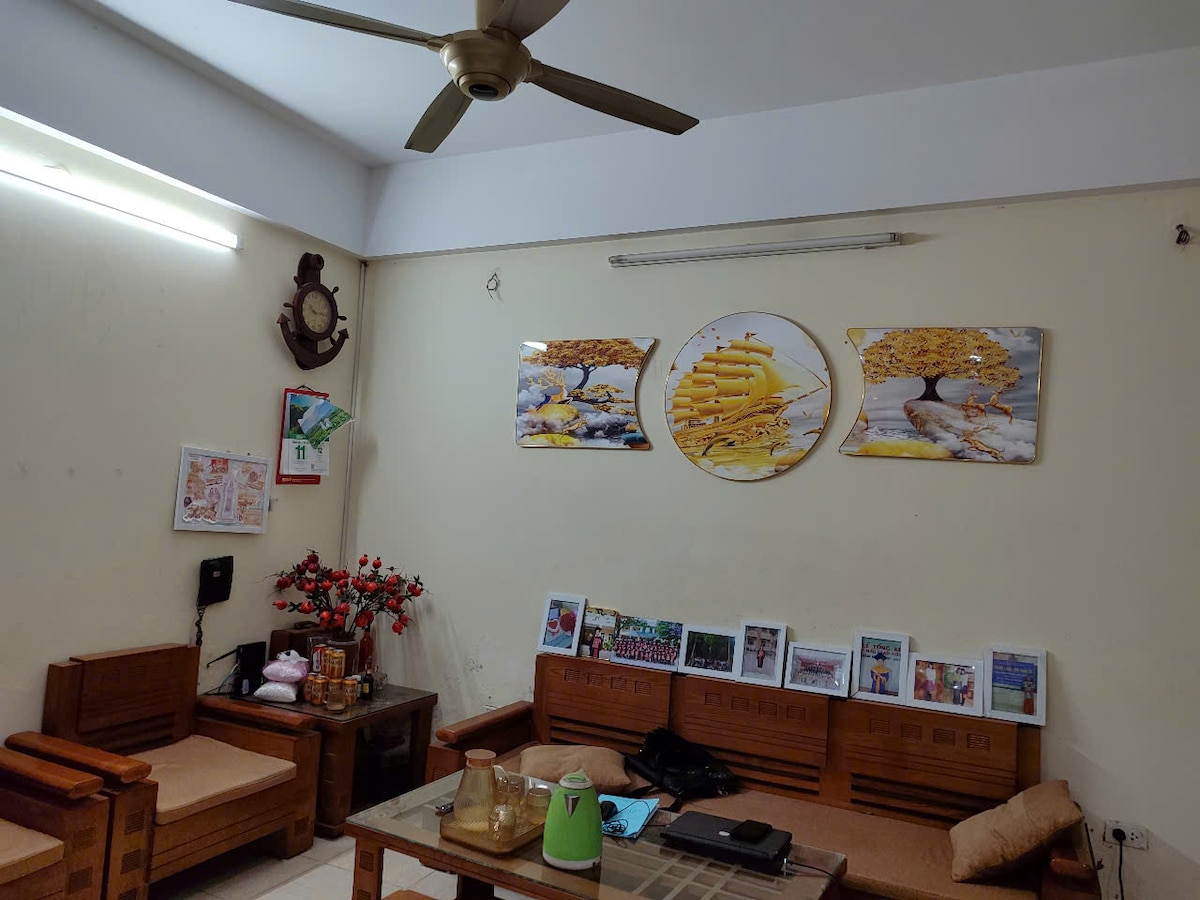 A comfortable living area features wooden seating arrangements on either side of a low table. Colorful wall artwork depicting trees adorns the beige walls. A ceiling fan is visible above, and small decorative items are arranged thoughtfully on the table.