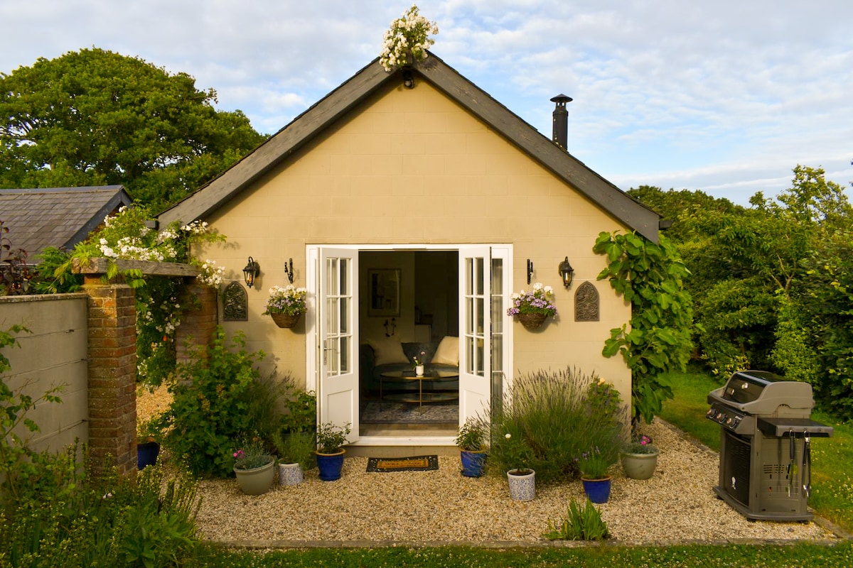 The exterior of a boutique barn conversion is visible, featuring double French doors framed by potted flowers. A BBQ grill is positioned nearby, and a gravel pathway leads to the entrance, surrounded by greenery and additional planters.