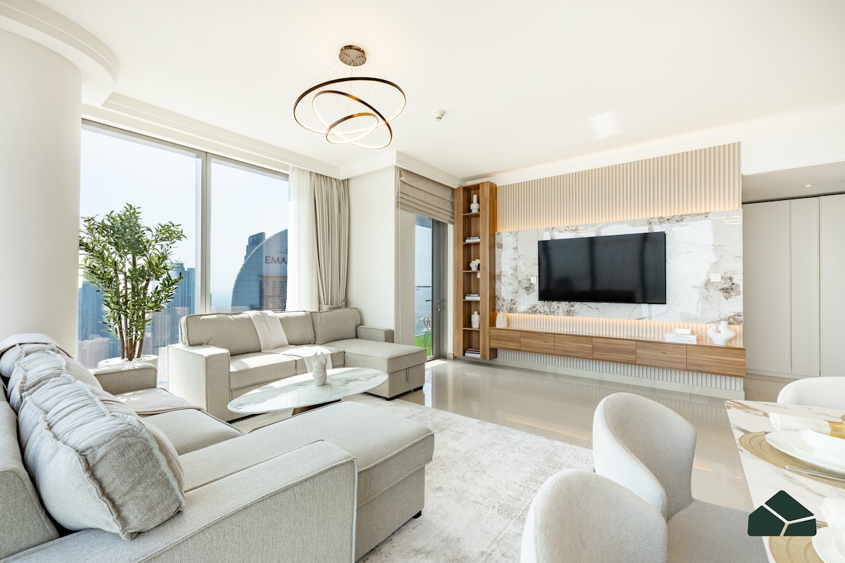 A spacious living room is displayed, featuring plush grey seating and a modern coffee table. Floor-to-ceiling windows provide expansive views of the city skyline. A large smart TV is mounted on a stylish wall, complemented by decorative shelving with curated items.