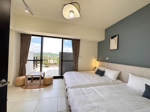 The sleeping area includes two spacious beds arranged along a light-colored wall. A small table is positioned between the beds, with a rug underneath. Floor-to-ceiling glass doors offer views of the outdoor balcony, enhancing natural light in the room.