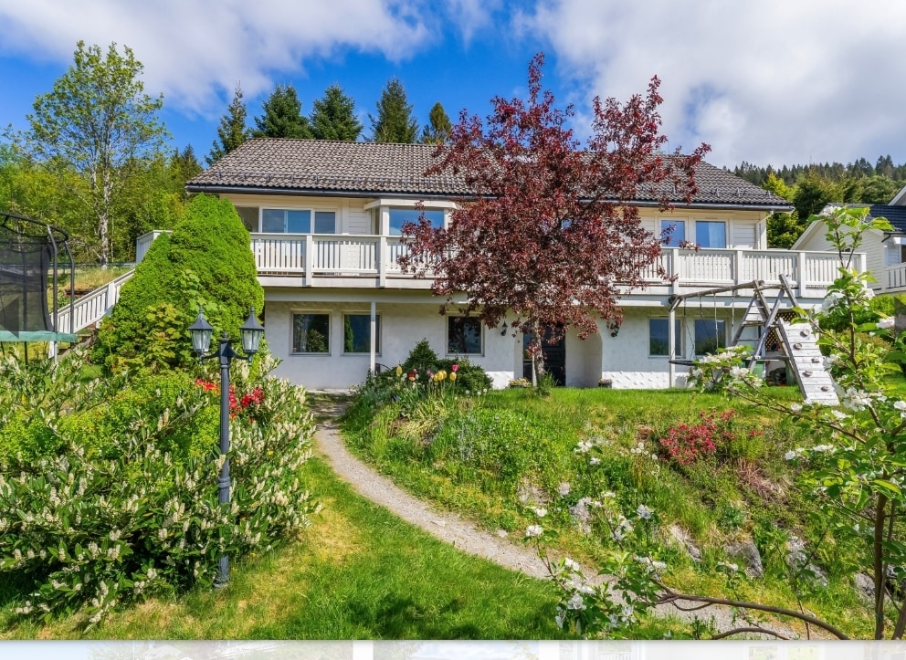 A two-story family home is set in a lush garden with vibrant flowers and greenery. A pathway leads to the entrance, flanked by well-maintained bushes. A large tree provides shade in the front yard, while a swing set is located on the right side.