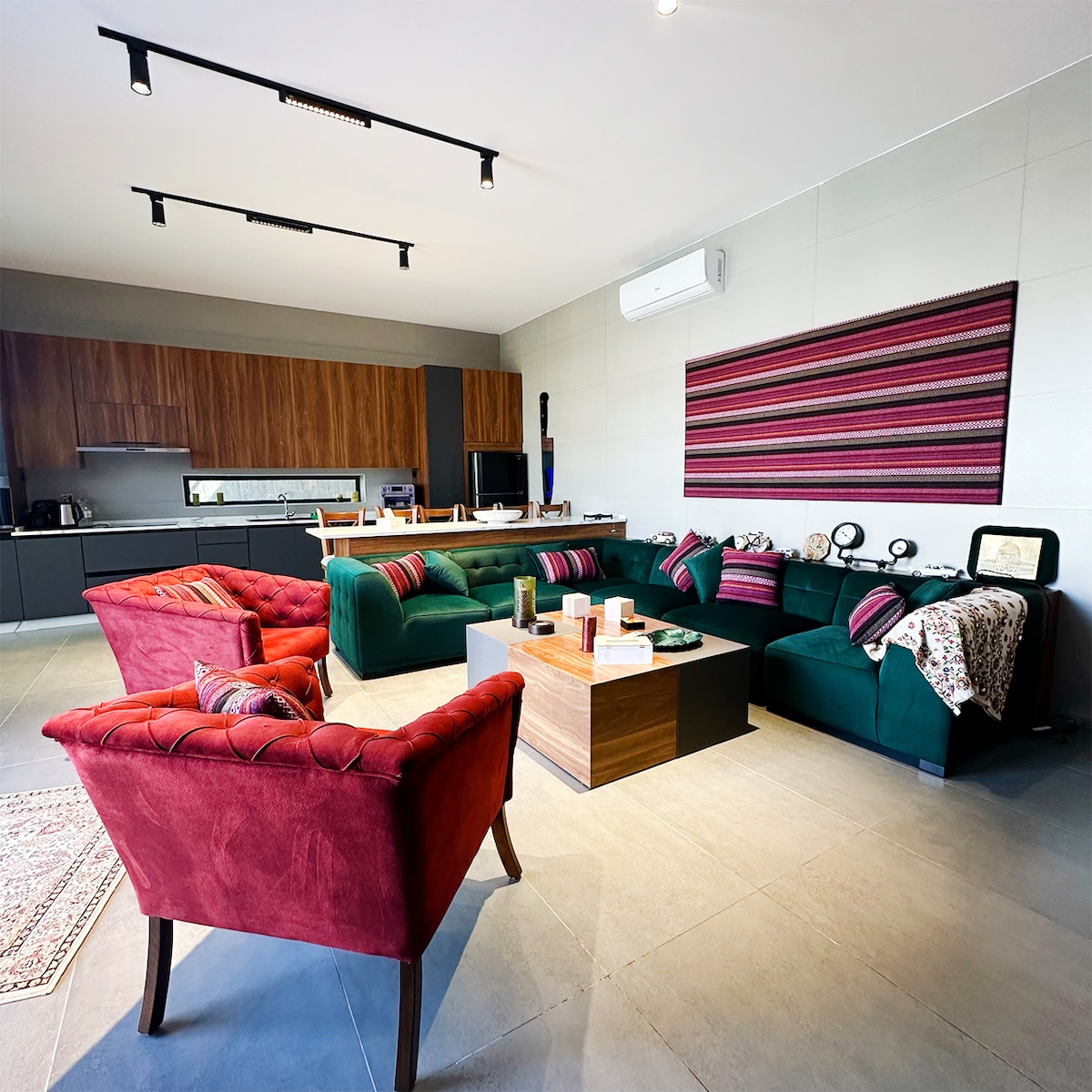 The living area features an emerald-green L-shaped sectional sofa, two plush red armchairs, and a large coffee table with local décor accents. The open-plan kitchen is visible in the background, with sleek cabinetry and modern appliances, creating a bright and inviting atmosphere.