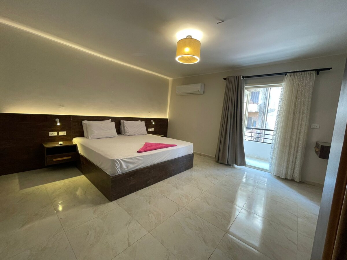 A bright and spacious room features a large bed with white linens and a pink throw. Soft lighting is provided by a pendant lamp. A sliding glass door leads to a private balcony, while light-colored curtains frame the window, allowing natural light to fill the space.